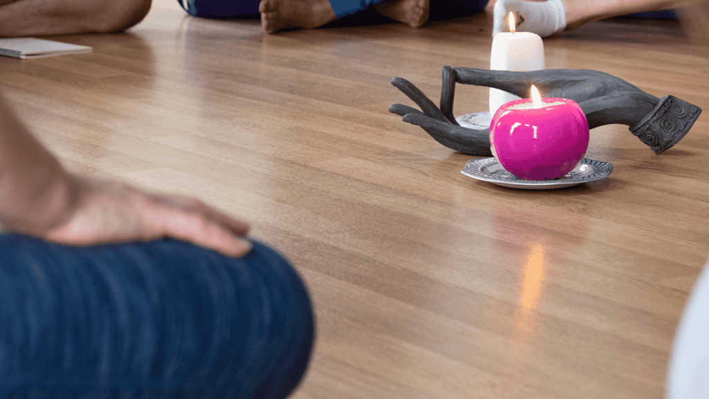 A picture of people on a yoga retreat sitting around a small ornament of a hand that's holding a Audra as well as two candles.